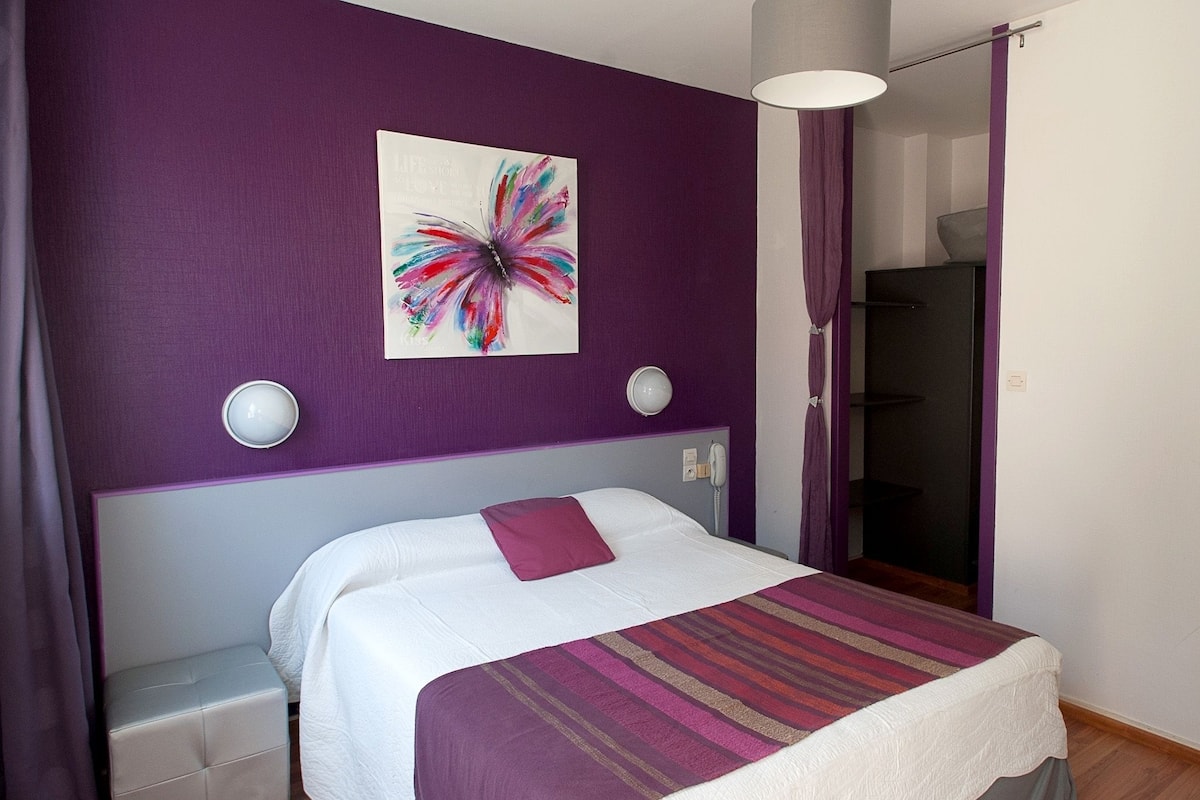 Une chambre à coucher moderne avec un grand lit, des murs violets, une œuvre d’art colorée et une table de chevet avec une lampe.