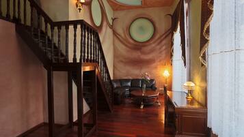 Imponente scalinata con ringhiera in legno che conduce a un soffitto alto decorato con motivi circolari. In primo piano, un divano in pelle e un tavolino rotondo, con una lampada su un comodino.