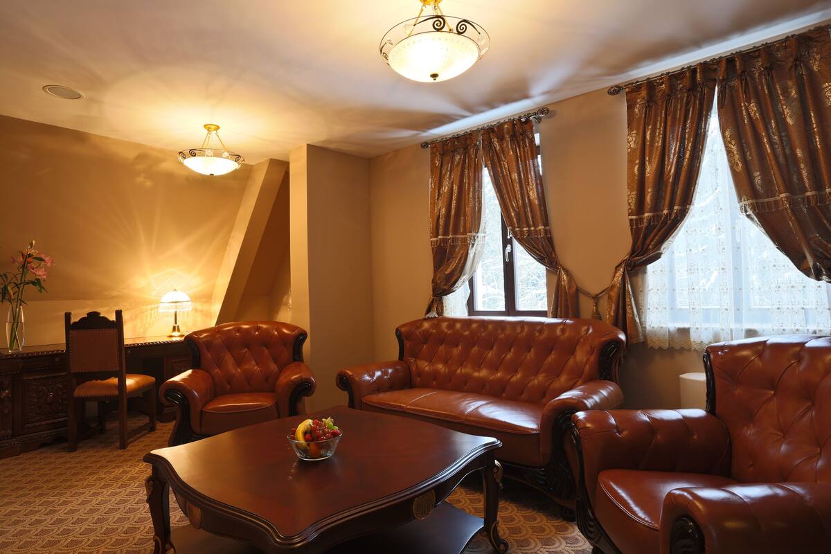 A room with a leather sofa, two armchairs, a wooden coffee table with fruit, a desk with a lamp, and a chandelier.