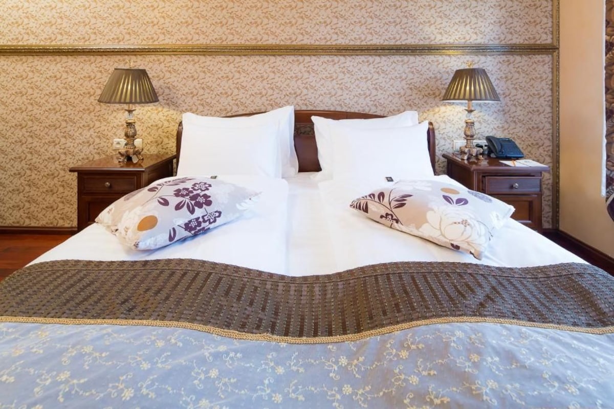 Un lit bien fait, agrémenté d’oreillers à motifs floraux et recouvert d’une couverture marron, avec deux tables de chevet de chaque côté, sur lesquelles reposent des lampes et un téléphone.