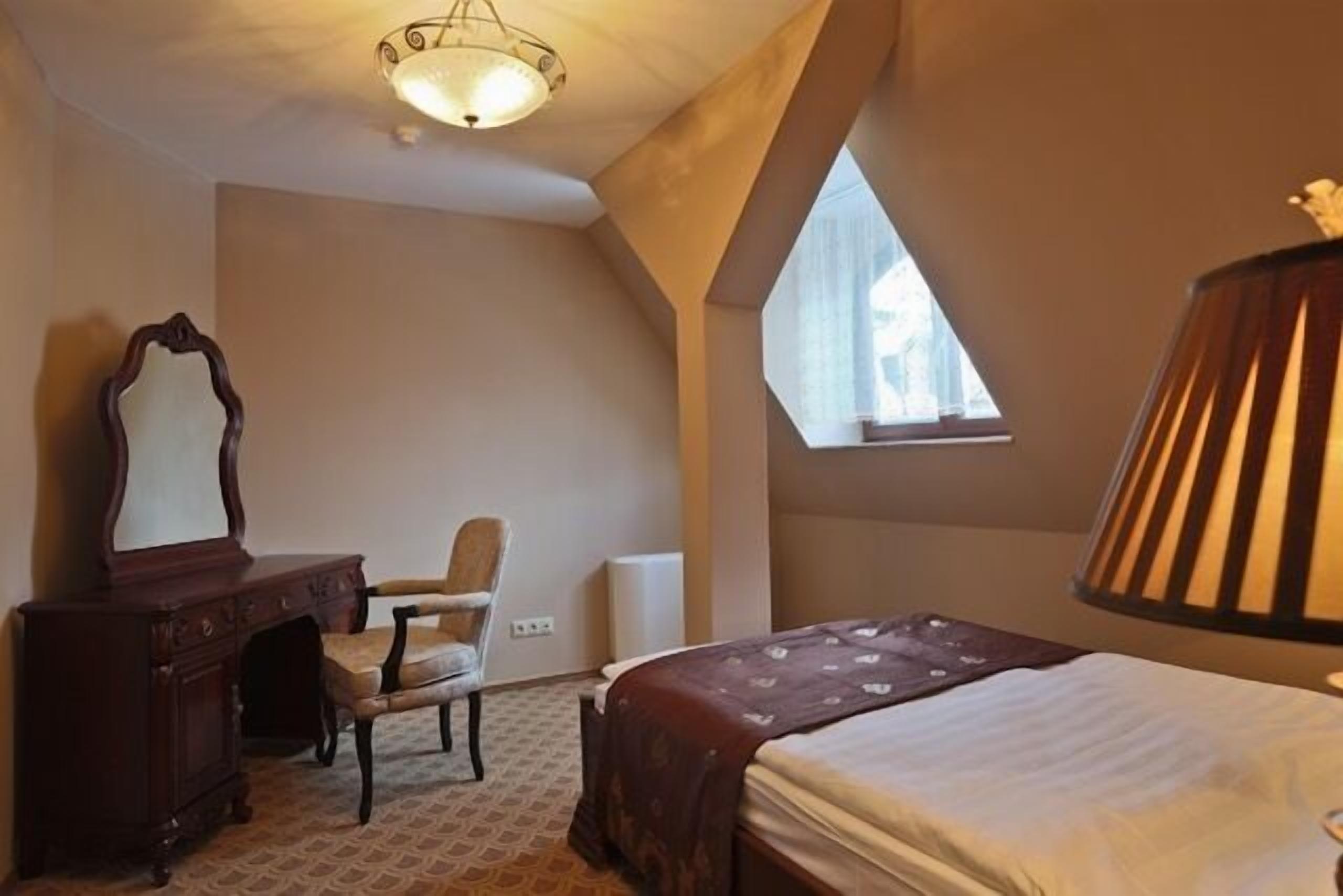 Un dormitorio con cama, una silla, tocador, lámpara y ventana.
