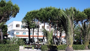 Un edificio bianco con numerosi balconi e un tetto in tegole rosse, immerso nel verde e circondato da palme.