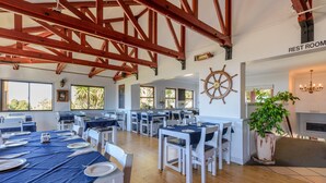 A spacious dining area with multiple tables set for service, a visible rest room sign, and a large potted plant. - Castleton (Plettenberg Bay)
