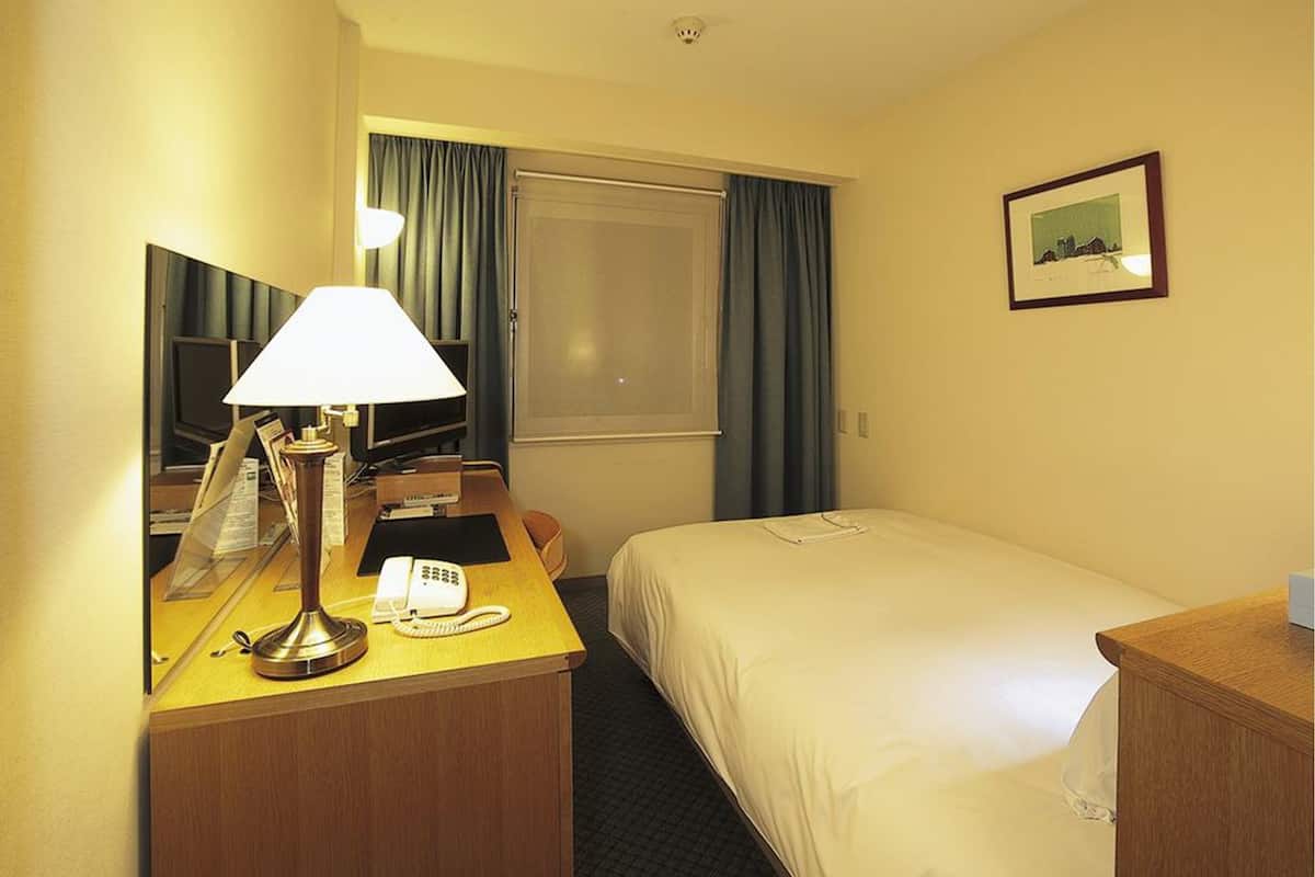 Ein Hotelzimmer mit einem Bett, einem Schreibtisch mit Lampe, einem Telefon und einem Fenster mit Vorhängen.
