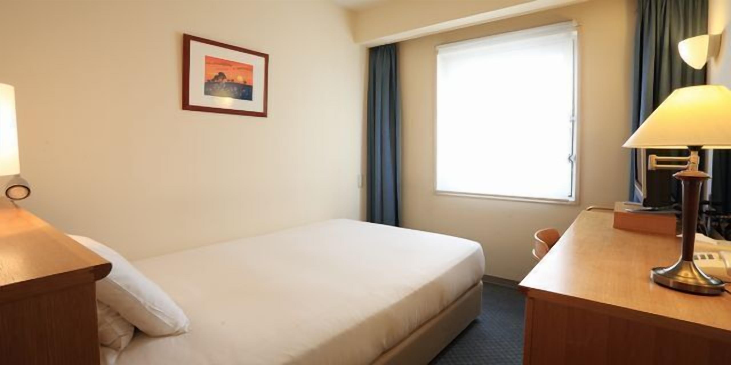 Une chambre d’hôtel avec un lit, un bureau doté d’une lampe, un tableau au mur et une fenêtre avec des rideaux.