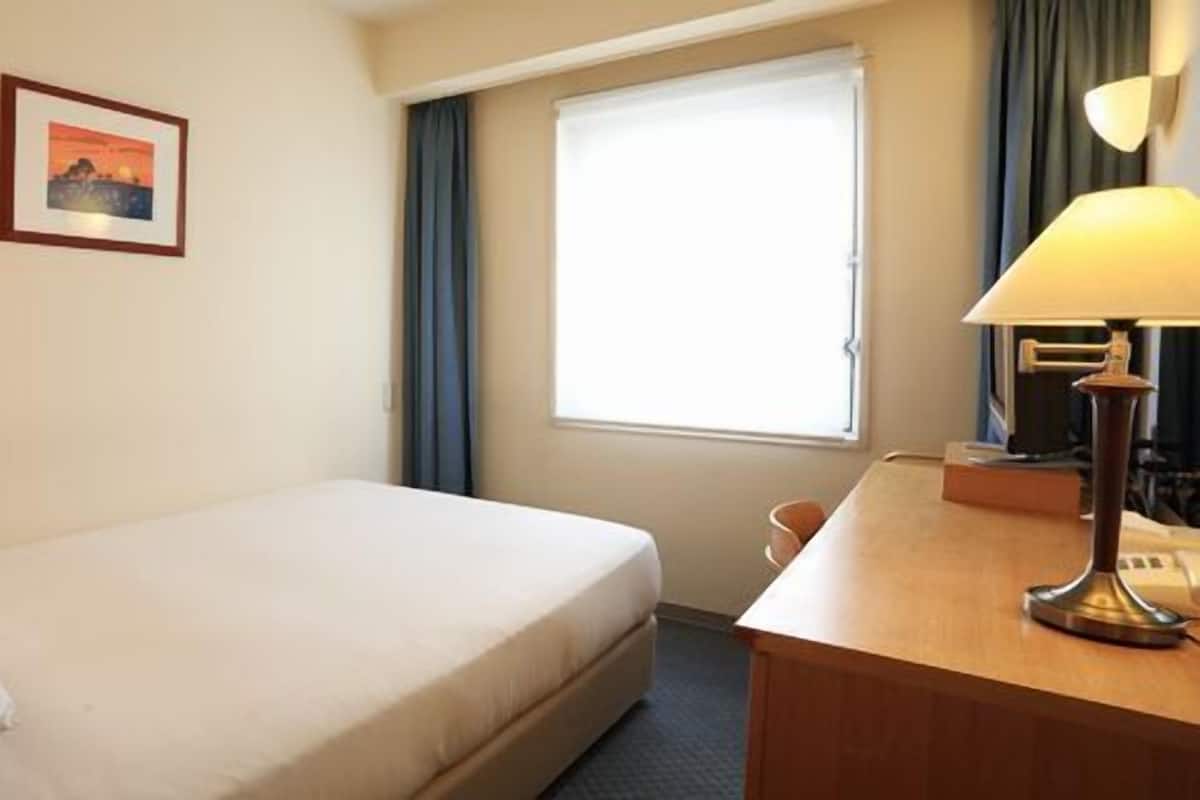 Ein Hotelzimmer mit Bett, Schreibtisch mit Lampe, einem Bild an der Wand und einem Fenster mit Vorhängen.