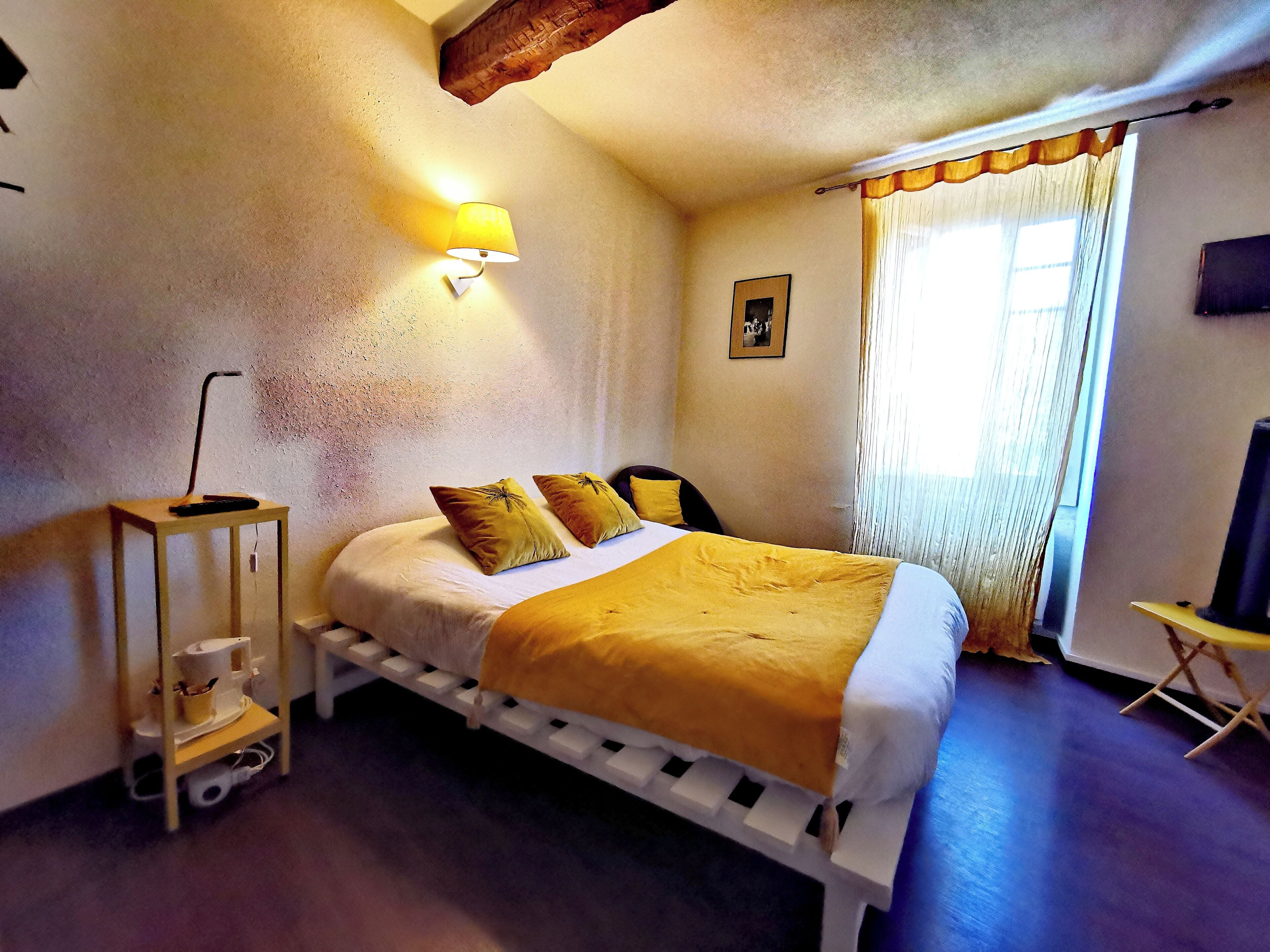 Un dormitorio con cama, una mesita de noche con lámpara, una silla, televisor y ventana con cortinas.