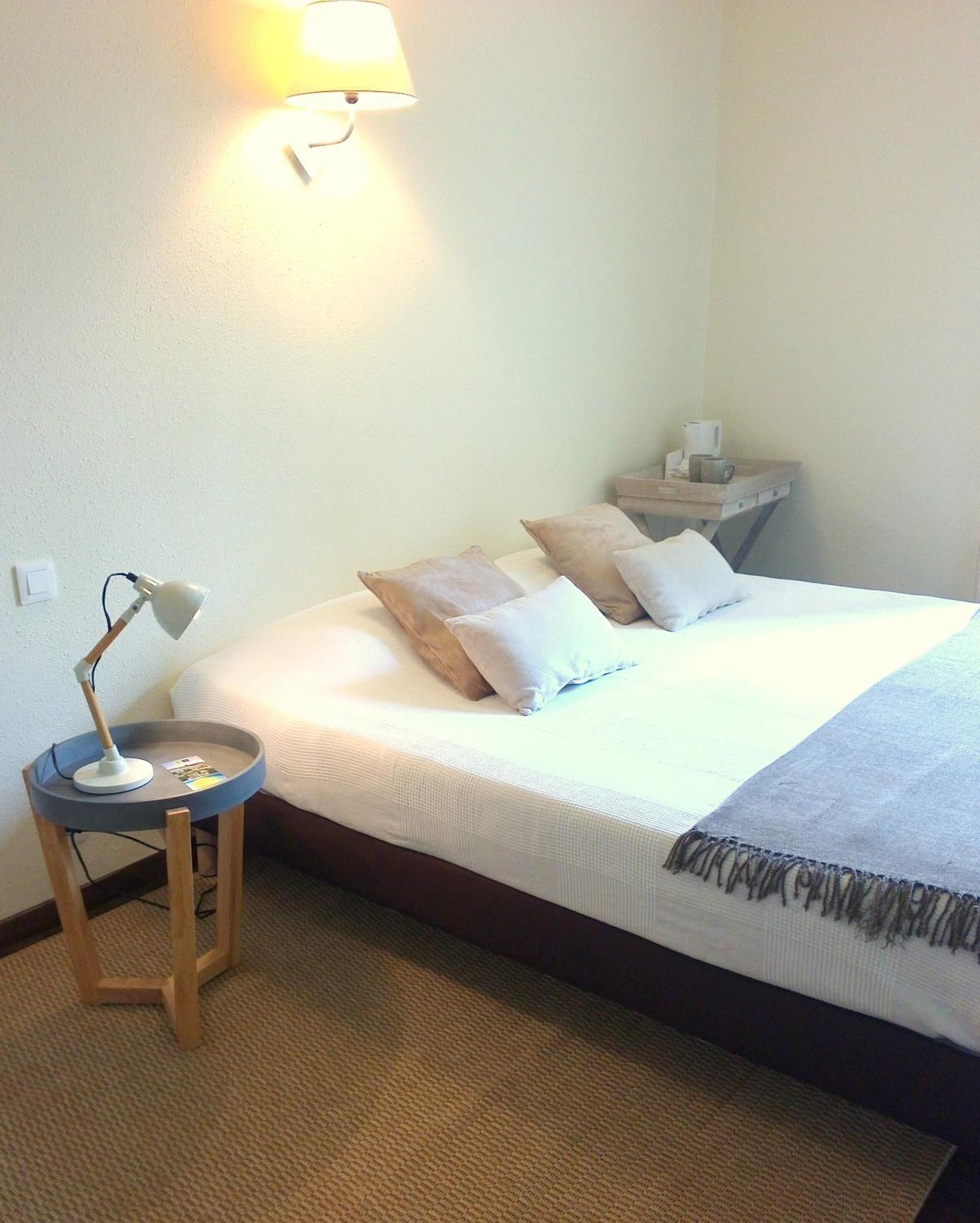 A neatly made bed with a white comforter and several pillows, a small bedside table with a lamp, and a wall-mounted light fixture.