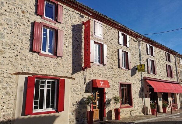A stone building with red shutters and a red awning, featuring a sign for 'La Barguillere' and a 'Slogis' sign. - Logis Auberge La Barguillère (Saint-Pierre-de-Riviere)