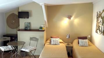 Una habitación pequeña y acogedora con dos camas individuales, una mesita con sillas, televisión y una lámpara fijada a la pared.