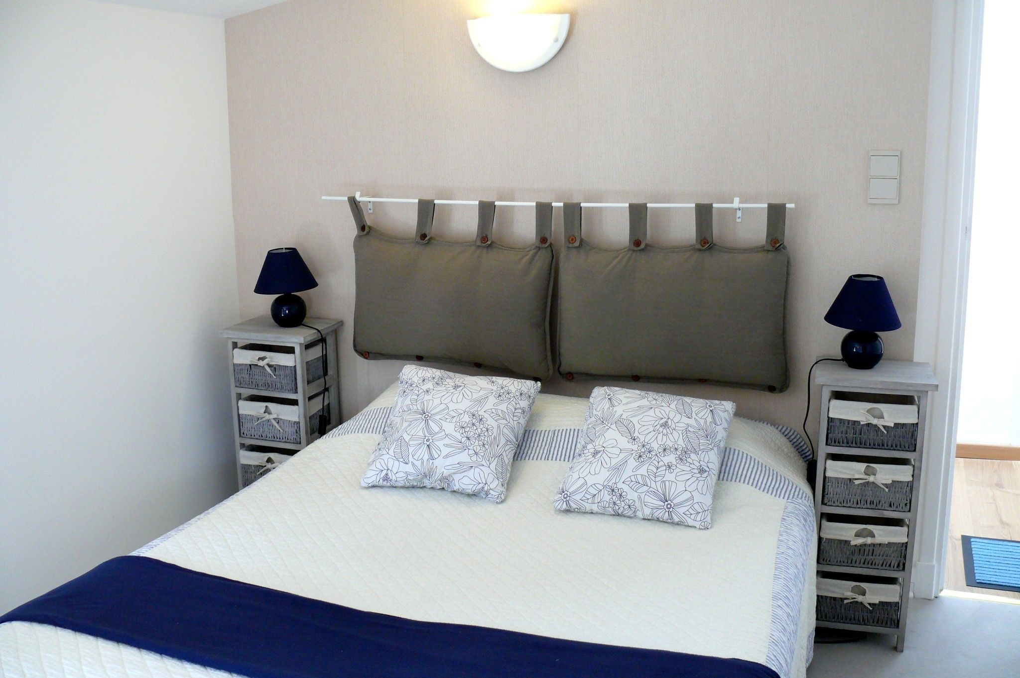 Una cama bien hecha con almohadas estampadas con motivos florales, una manta azul y un cabecero con cortinas. Hay dos mesitas de noche con lámparas y cajones.