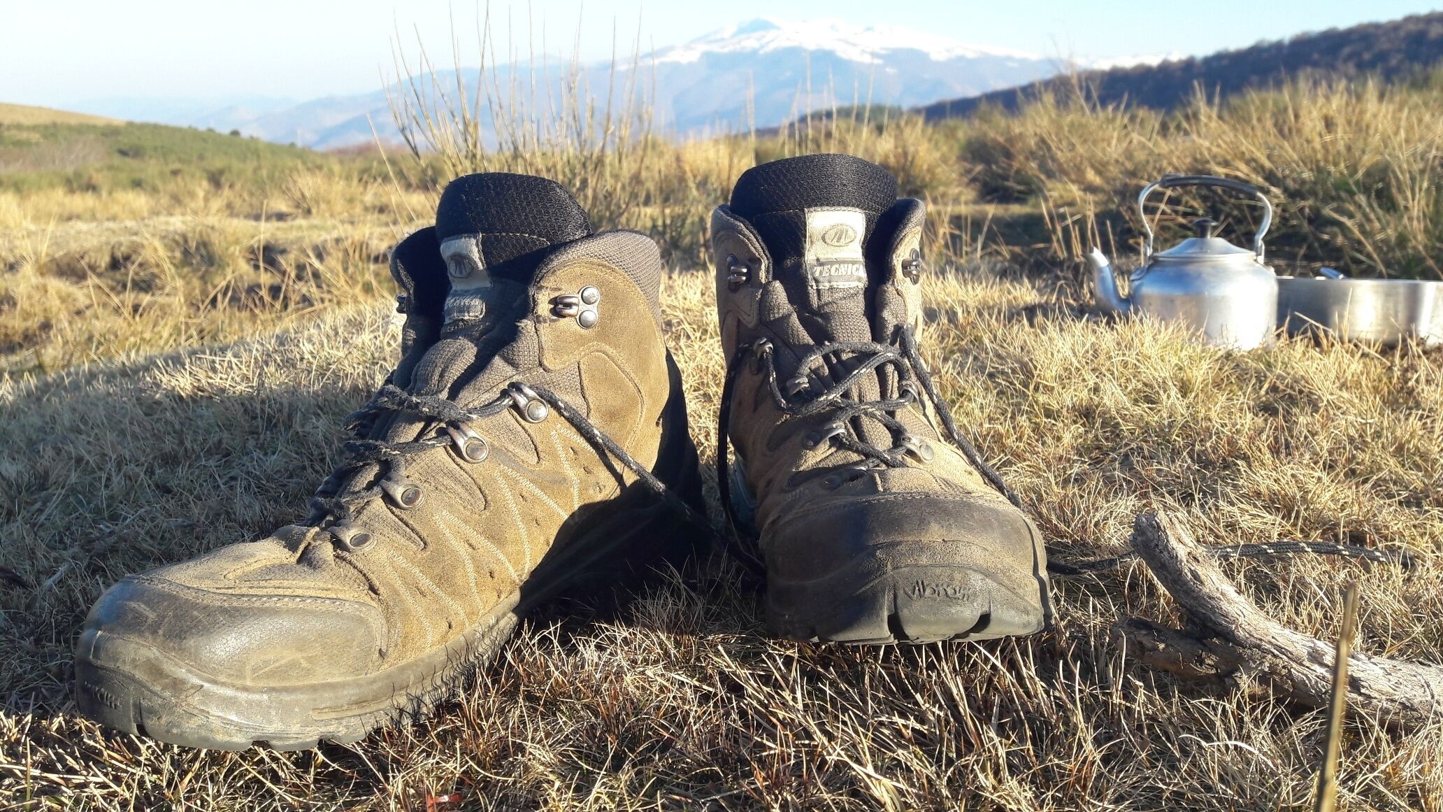 A pair of muddy hiking boots on dry grass with a metal kettle and a metal container in the background.