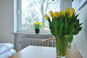 Ein Zimmer mit einem Bett, einem Tisch mit einer Vase voller gelber Tulpen und einem Fenster mit Blick auf Bäume und einer kleinen Topfpflanze.
