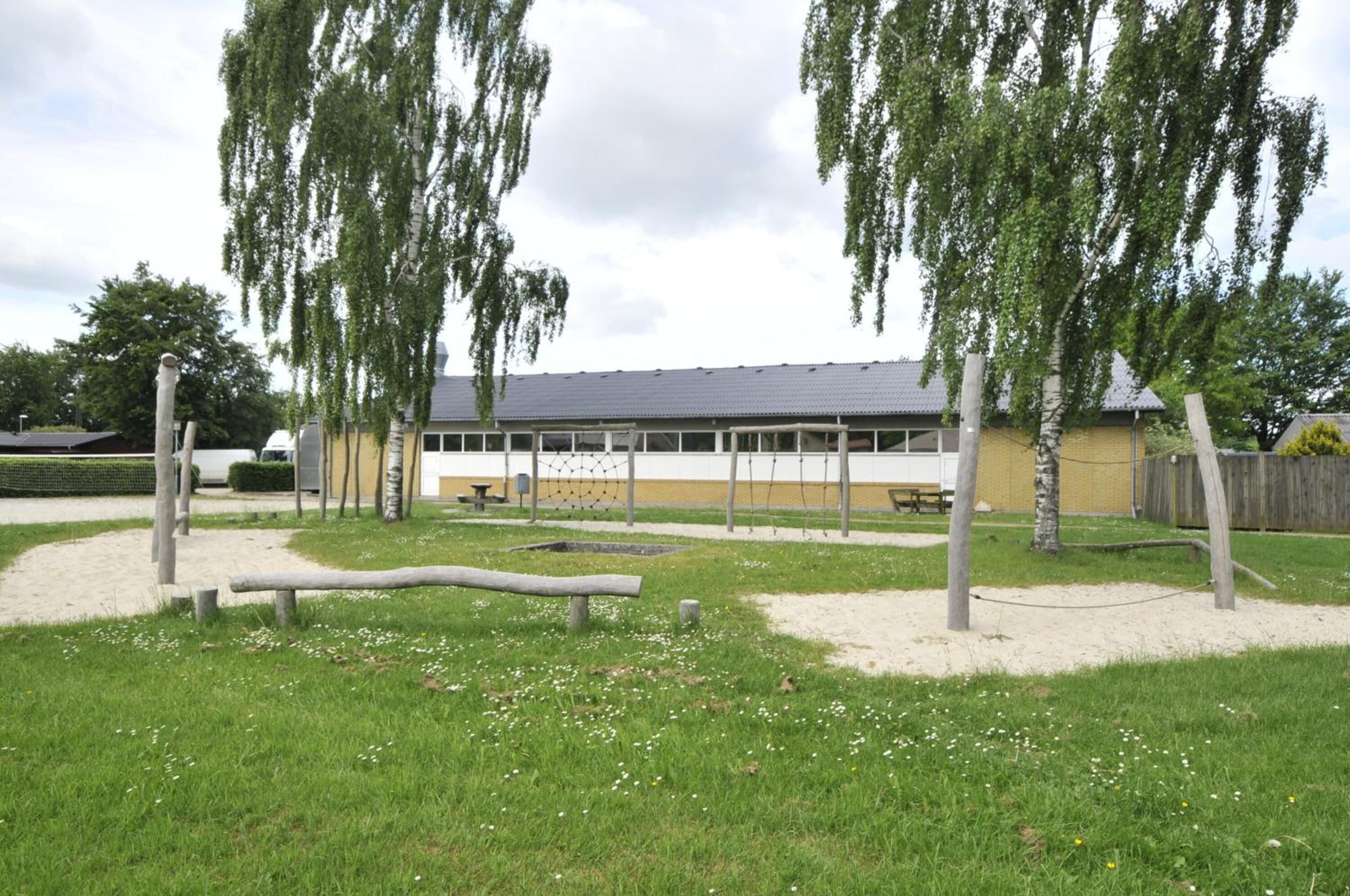A building with a flat roof and a yellow facade, surrounded by a grassy area with trees and a wooden fence.