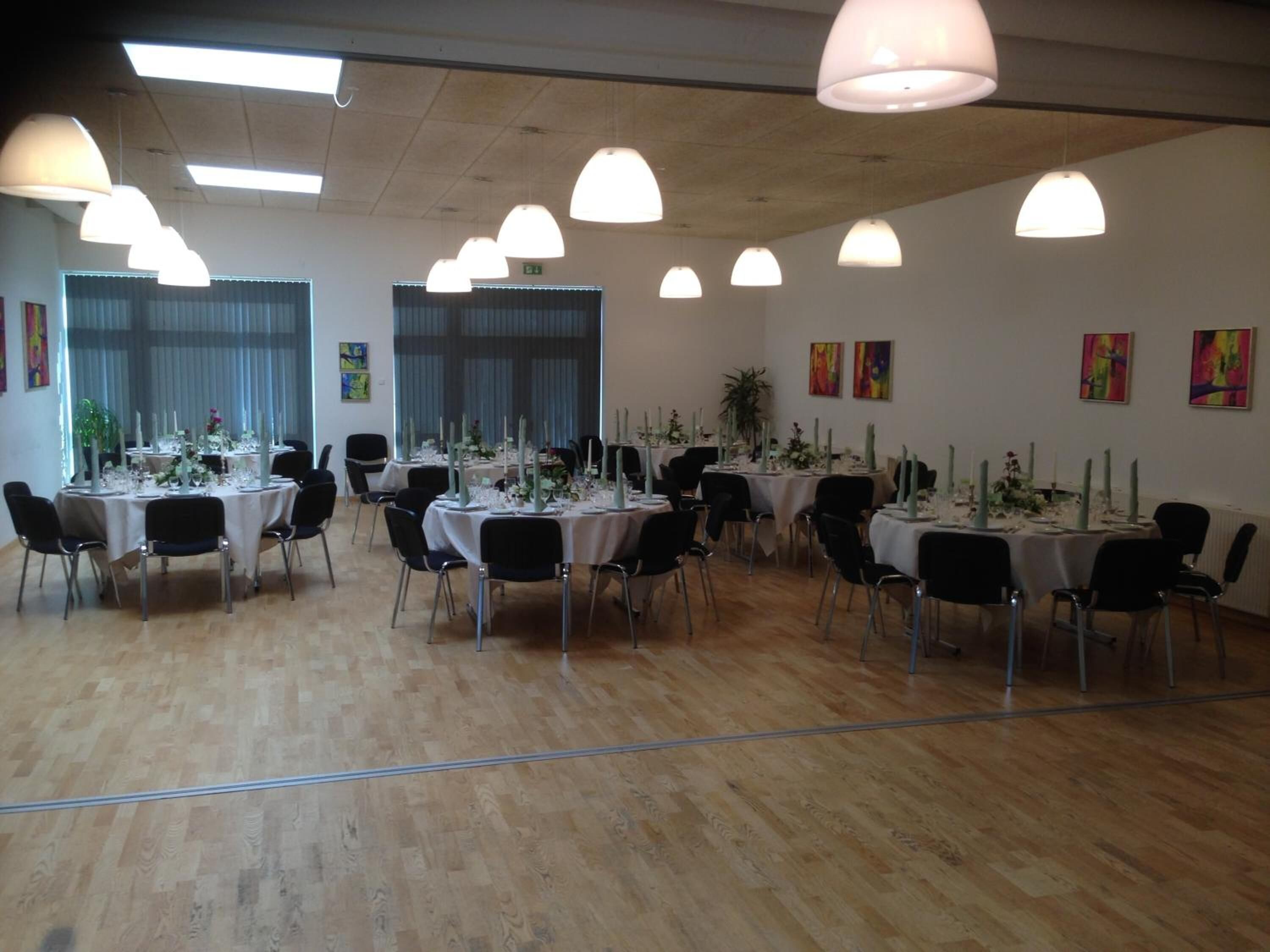 A spacious room with multiple round tables set for dining, each with white tablecloths and candle centerpieces.