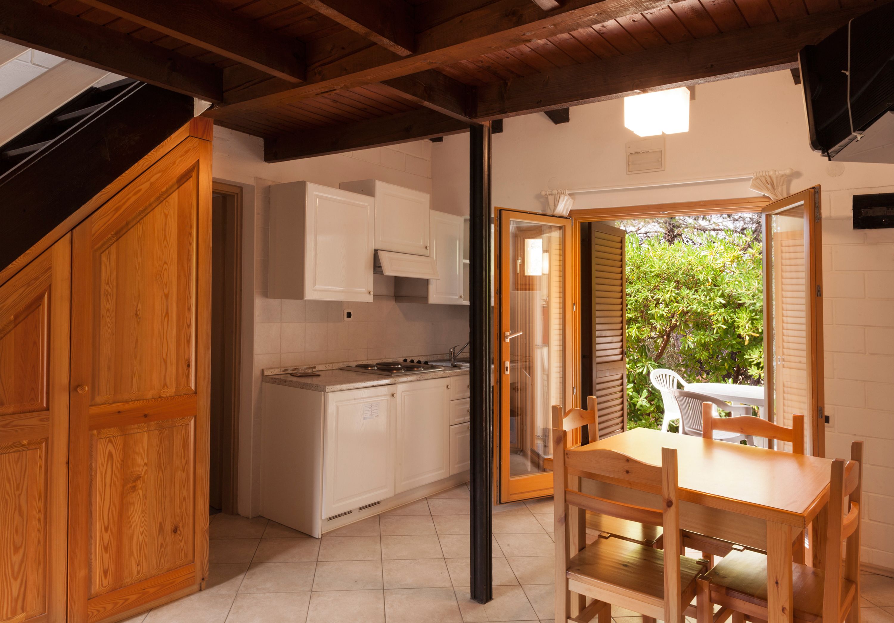 A kitchen with white cabinets, a wooden dining table, and chairs. There is a sliding glass door leading to an outdoor area with greenery.