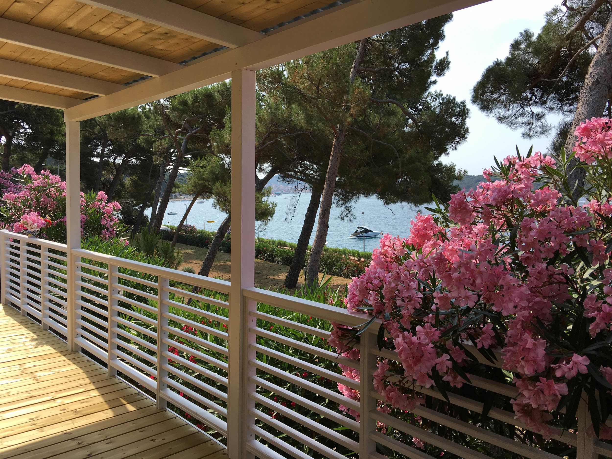 A wooden deck with white railings overlooking a lake with boats, surrounded by trees and pink flowers.