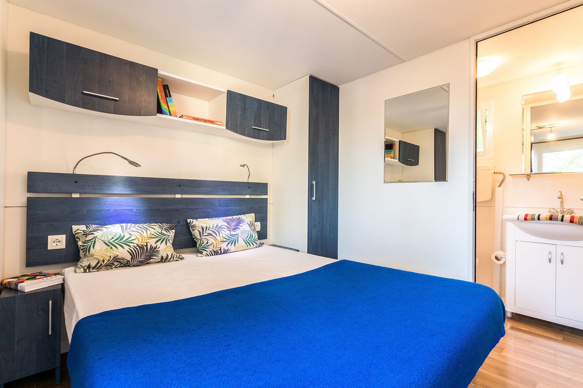 Ein modernes Schlafzimmer mit einem großen Bett, einem Nachttisch, einem Kleiderschrank und einem Badezimmer mit Waschbecken und Spiegel.