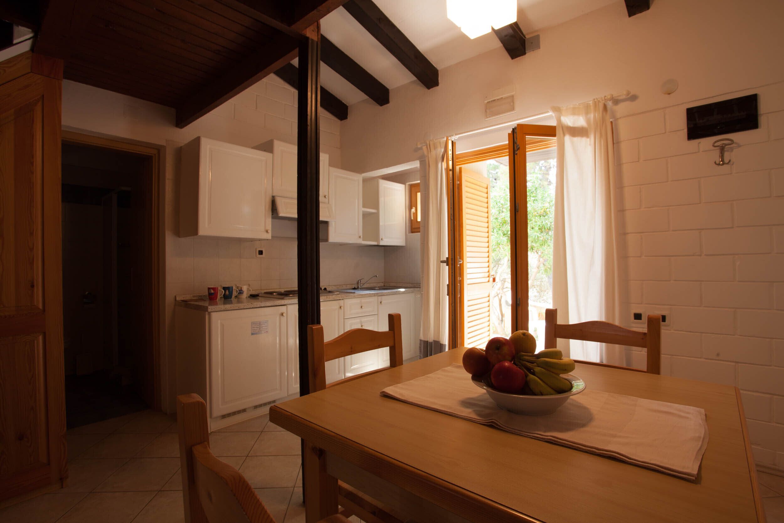 A kitchen with white cabinets, a wooden table with a bowl of fruit, and a window with curtains.