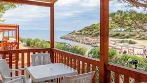 Eine Holzterrasse mit Tisch und Stühlen, mit Blick auf eine felsige Küstenlinie, Menschen und einen Strand.