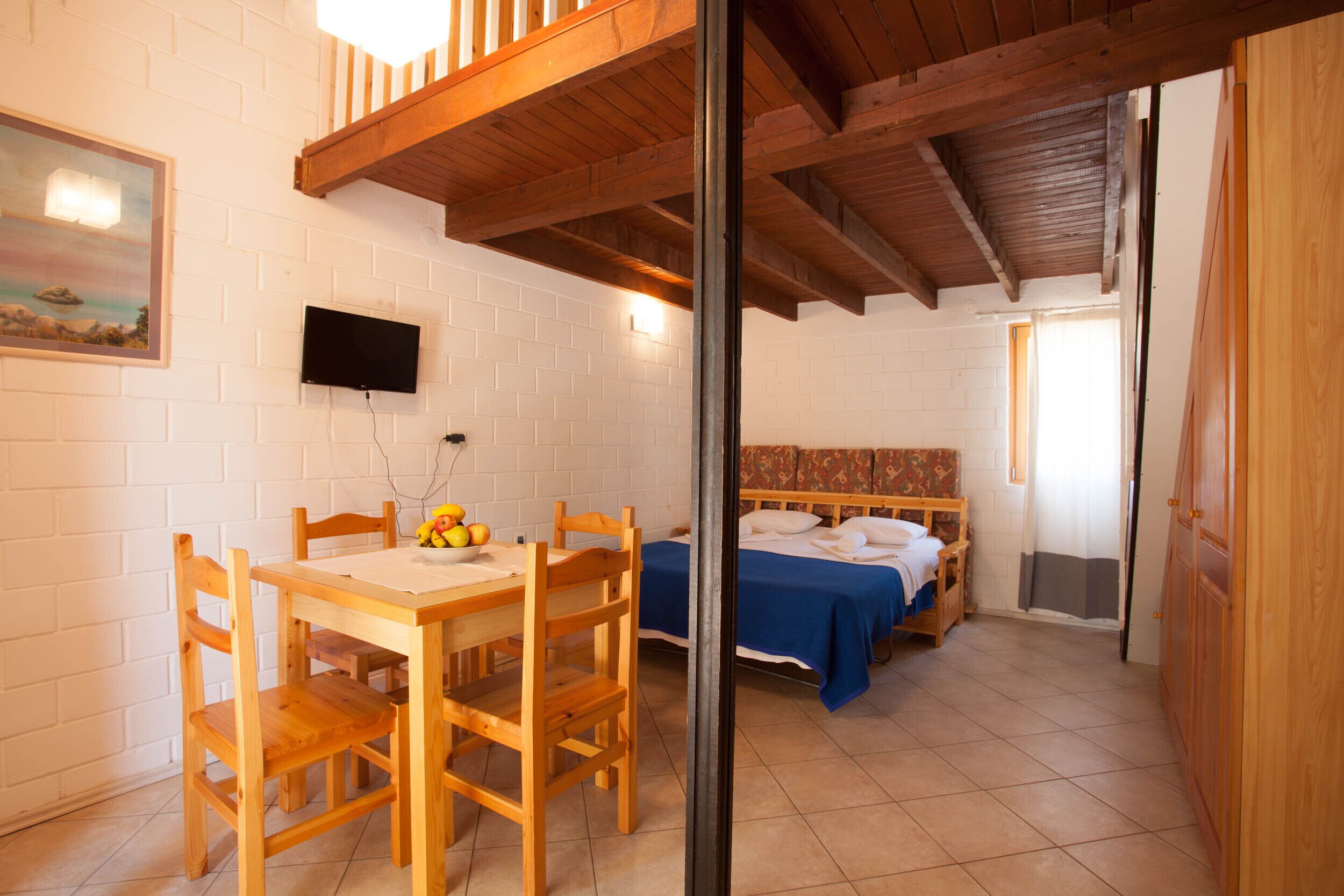 A room with a wooden dining table, chairs, a bed with a blue bedspread, and a TV mounted on the wall.