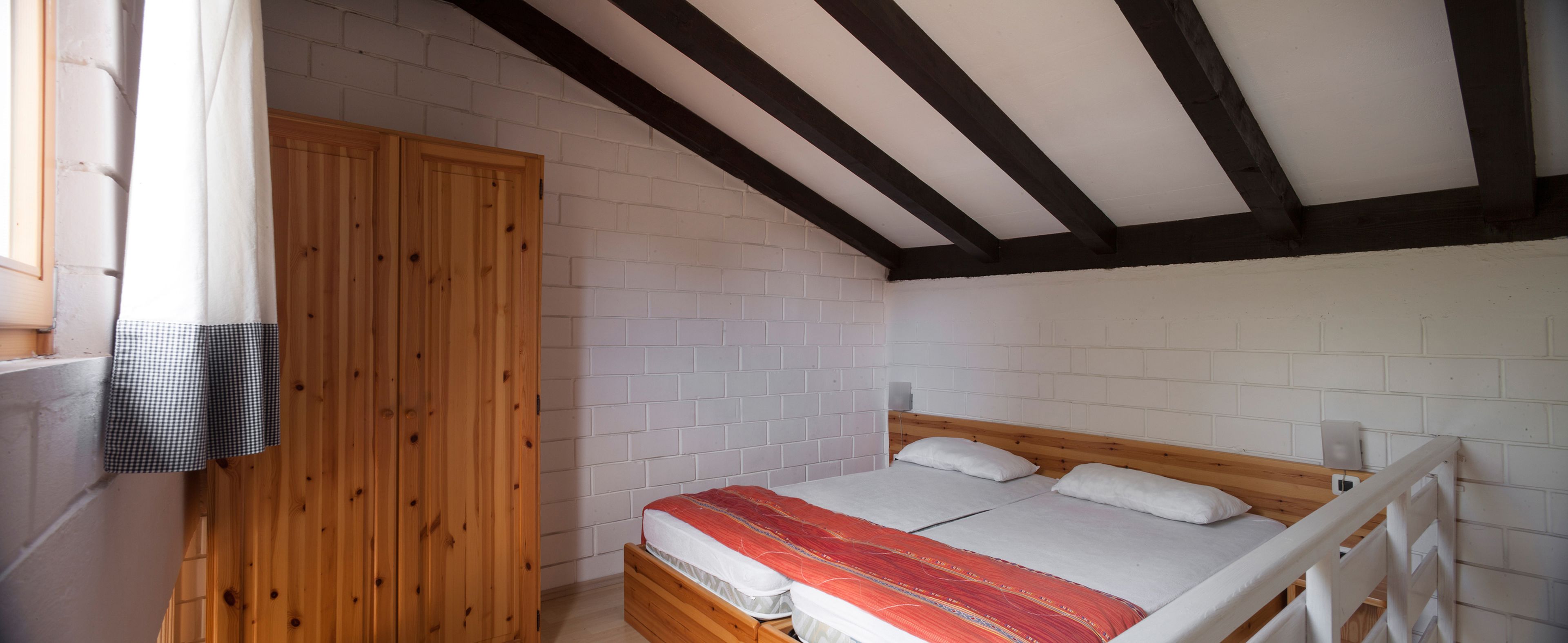 Et soveværelse med en træseng, hvide murstensvægge og et skråt loft med mørke bjælker.
