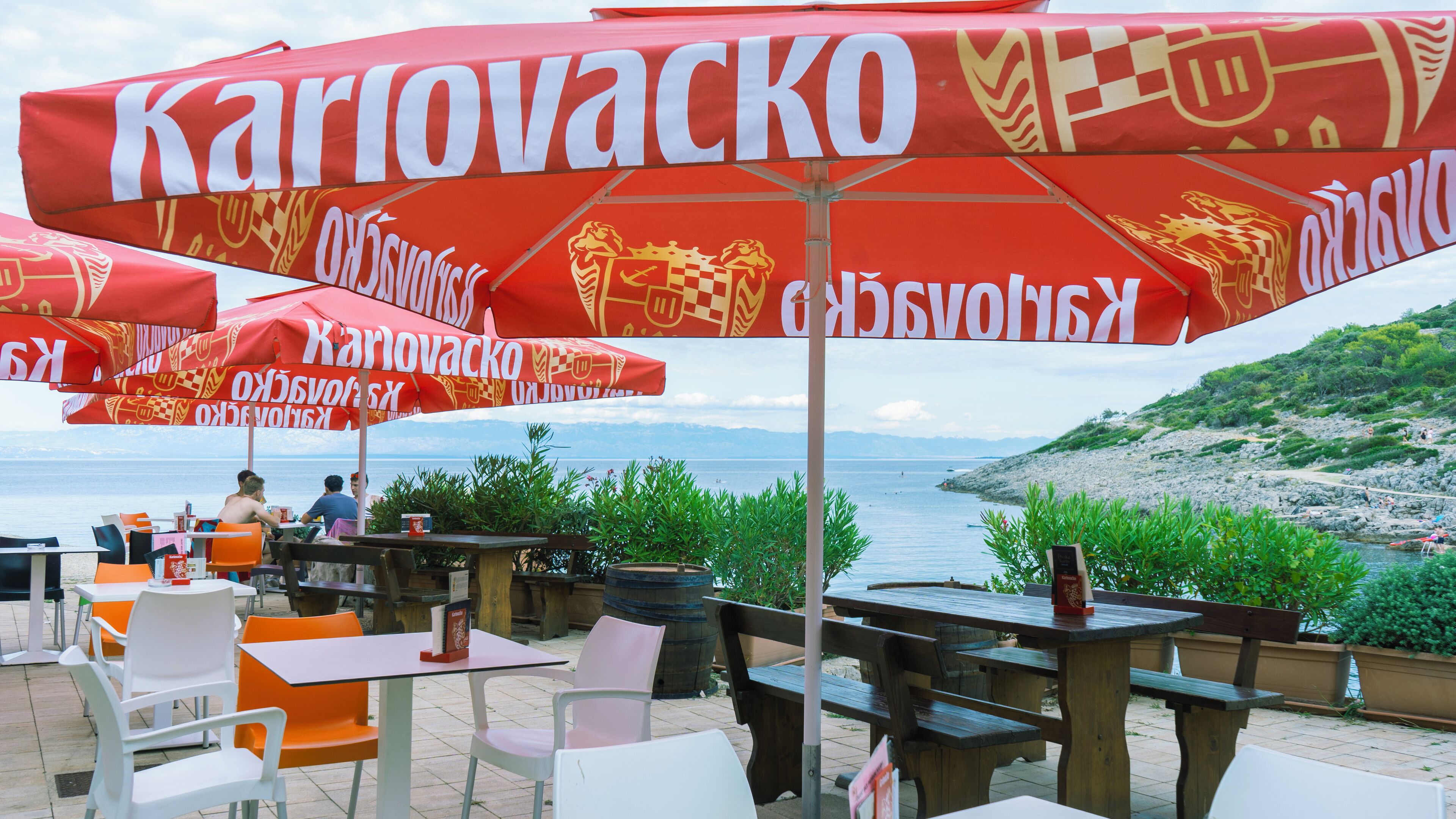 Εξωτερικός χώρος με τραπέζια και κόκκινες ομπρέλες, με θέα στη θάλασσα.
