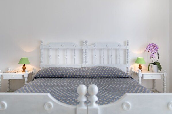 Cama bem arrumada com colcha azul e amarela estampada, cabeceira branca e mesas de cabeceira combinando com luminárias e uma orquídea em vaso.