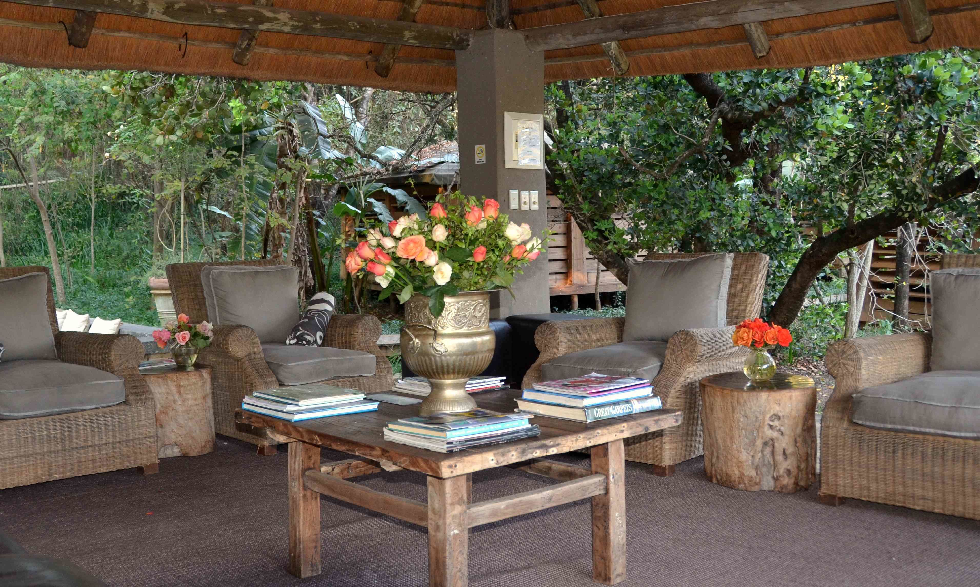 Un espacio al aire libre cubierto con mobiliario de mimbre, una mesa de madera con libros y un florero con flores.