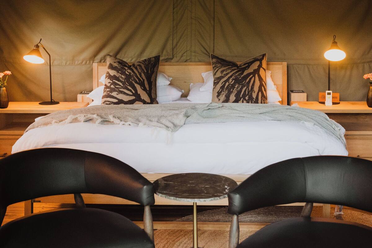 A tented accommodation room with a bed, two chairs, a table, and a ceiling fan.