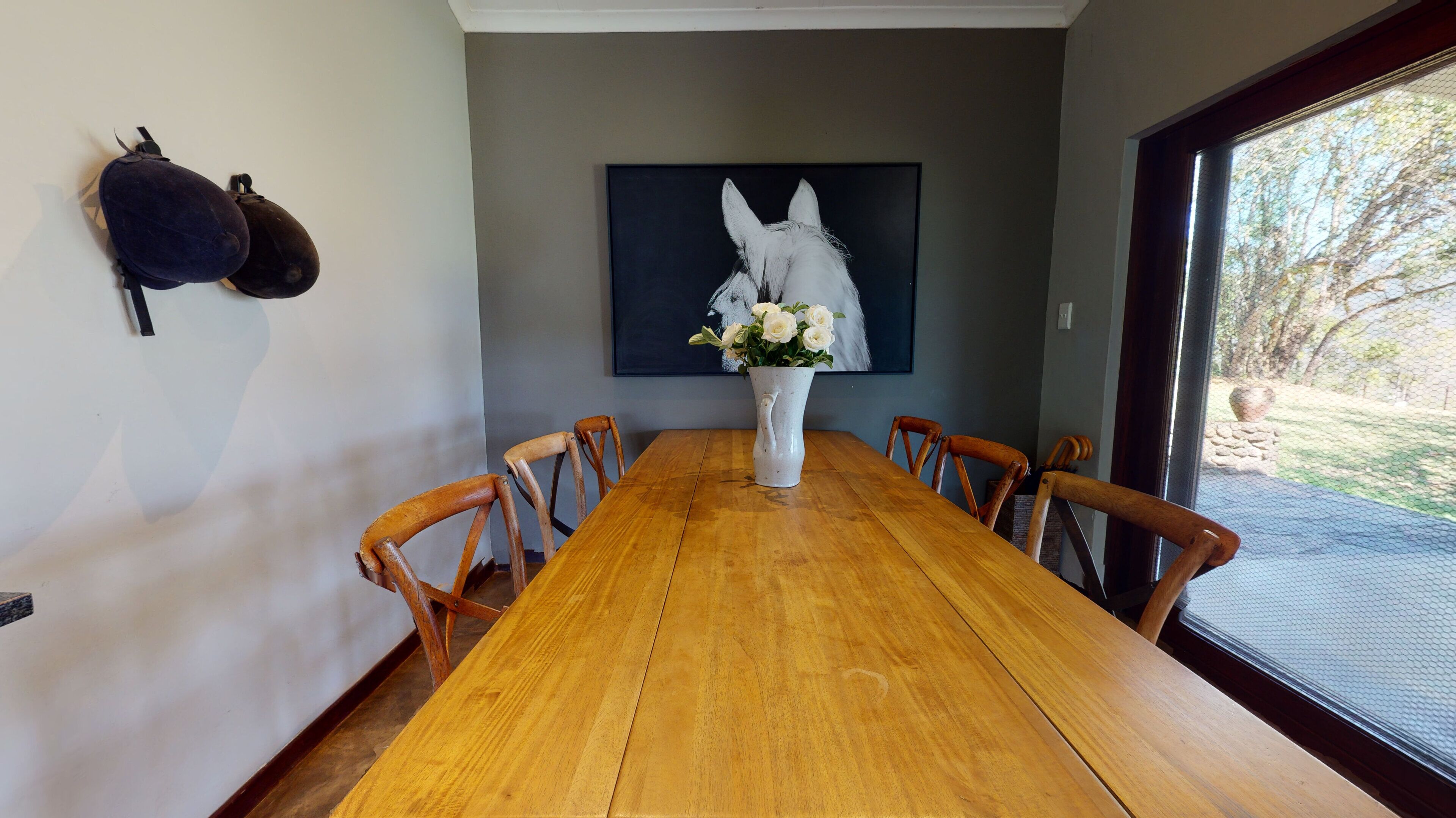 A dining room with a wooden table, six chairs, a vase with white flowers, and a large painting on the wall.