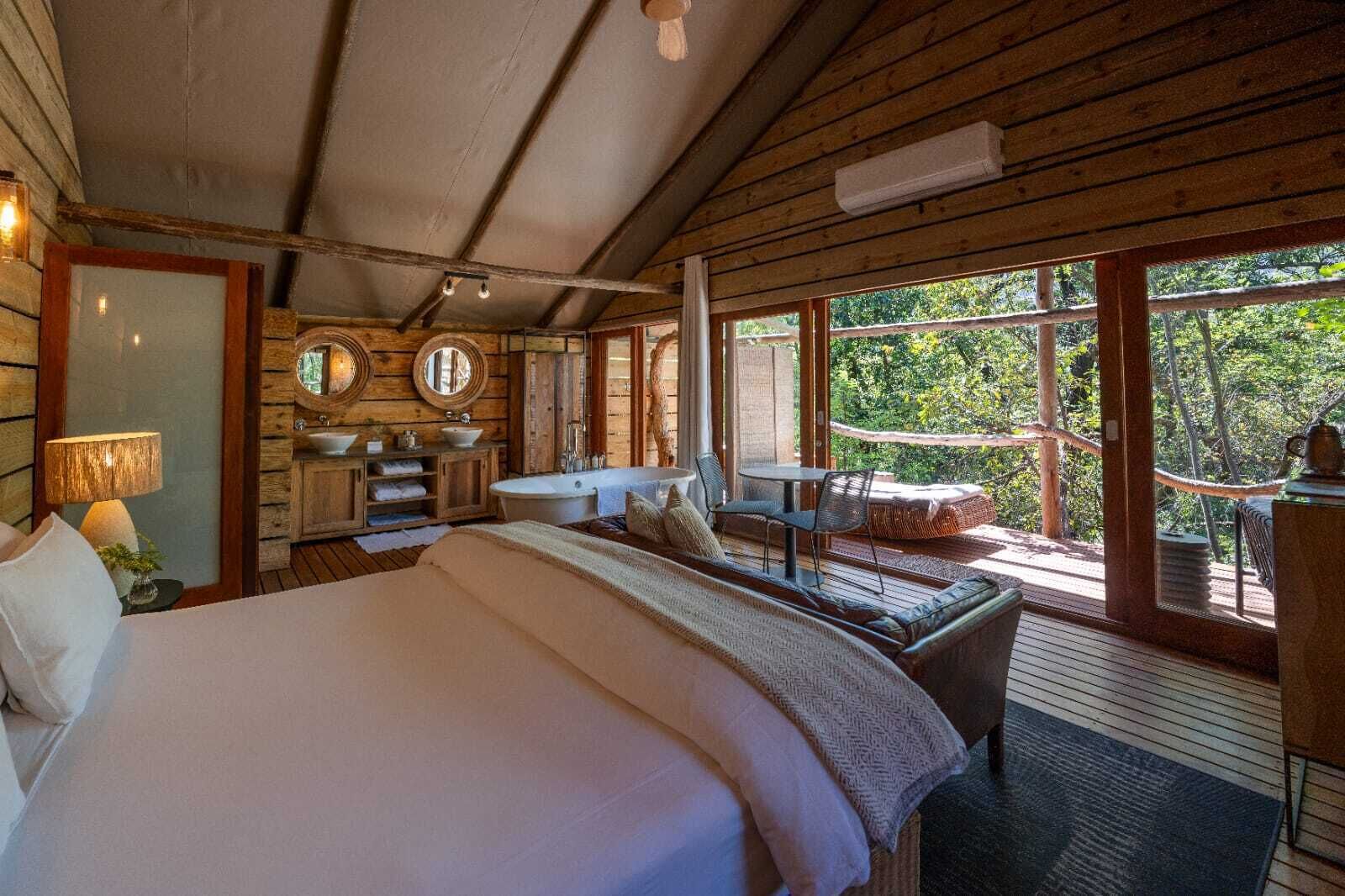 Une chambre dans un chalet en bois, avec un grand lit, une salle de bain dotée d’une baignoire et un balcon offrant une vue sur la forêt.