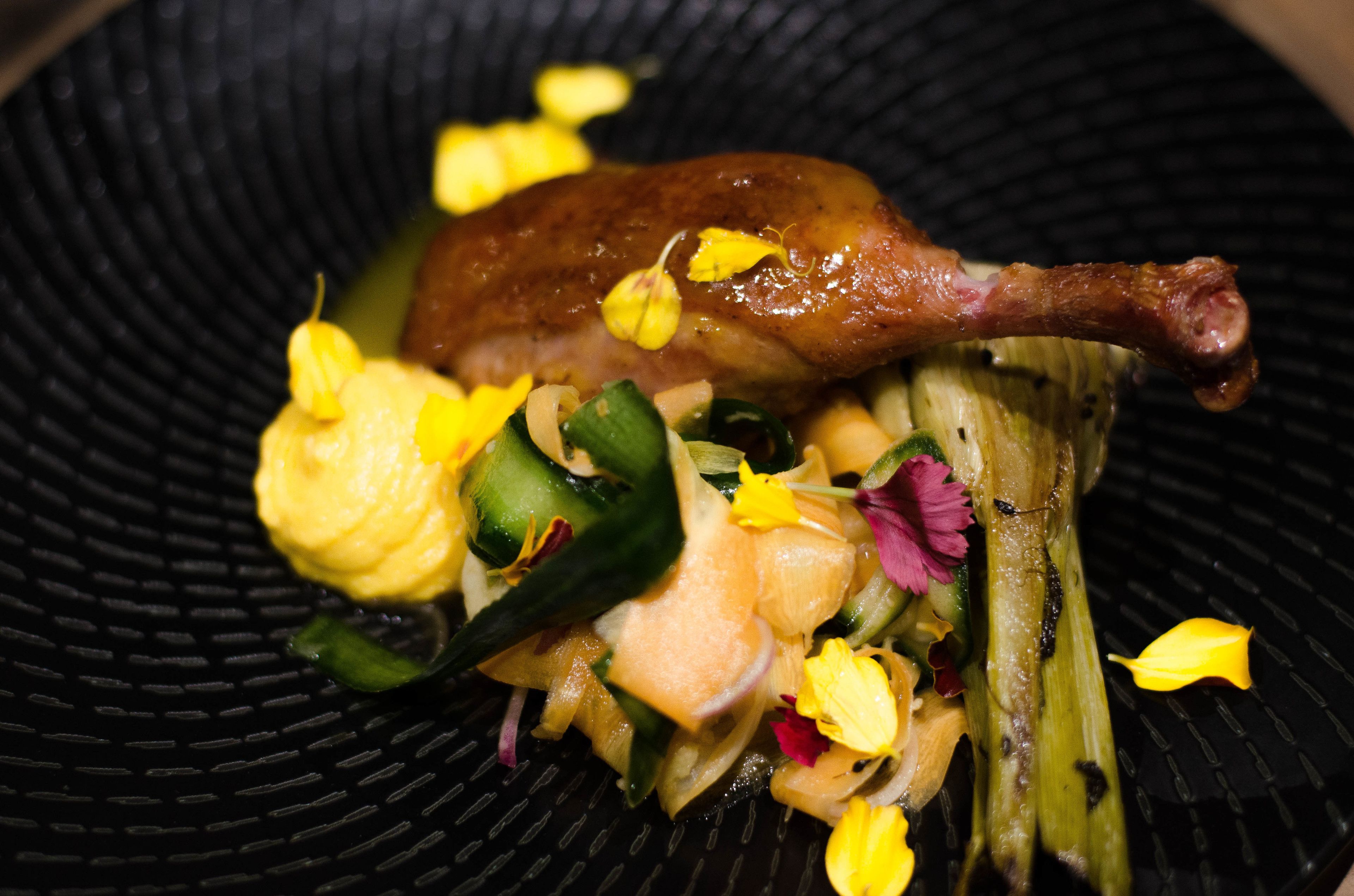 A dish with a roasted duck leg, vegetables, and flowers on a black plate.