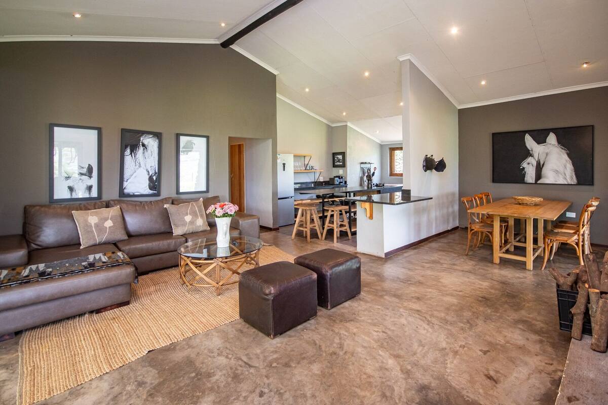 A modern living room with a sofa, coffee table, and ottoman, a dining area with a wooden table and chairs, and a kitchen in the background.