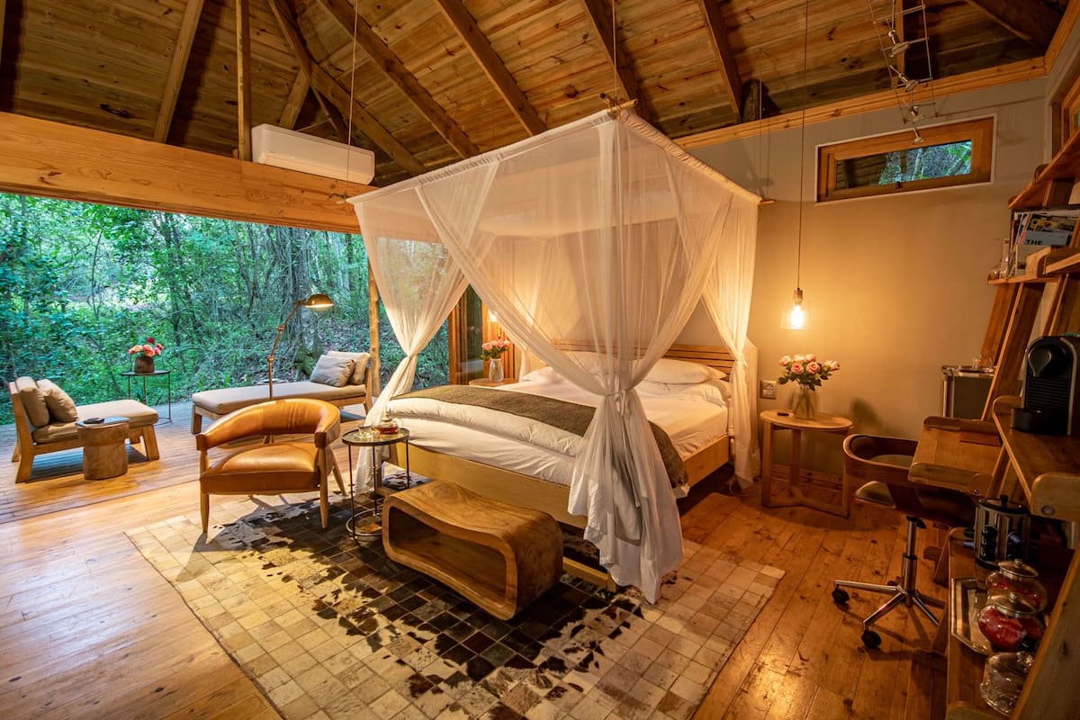 A cozy bedroom with a canopy bed, wooden furniture, and a view of the forest.