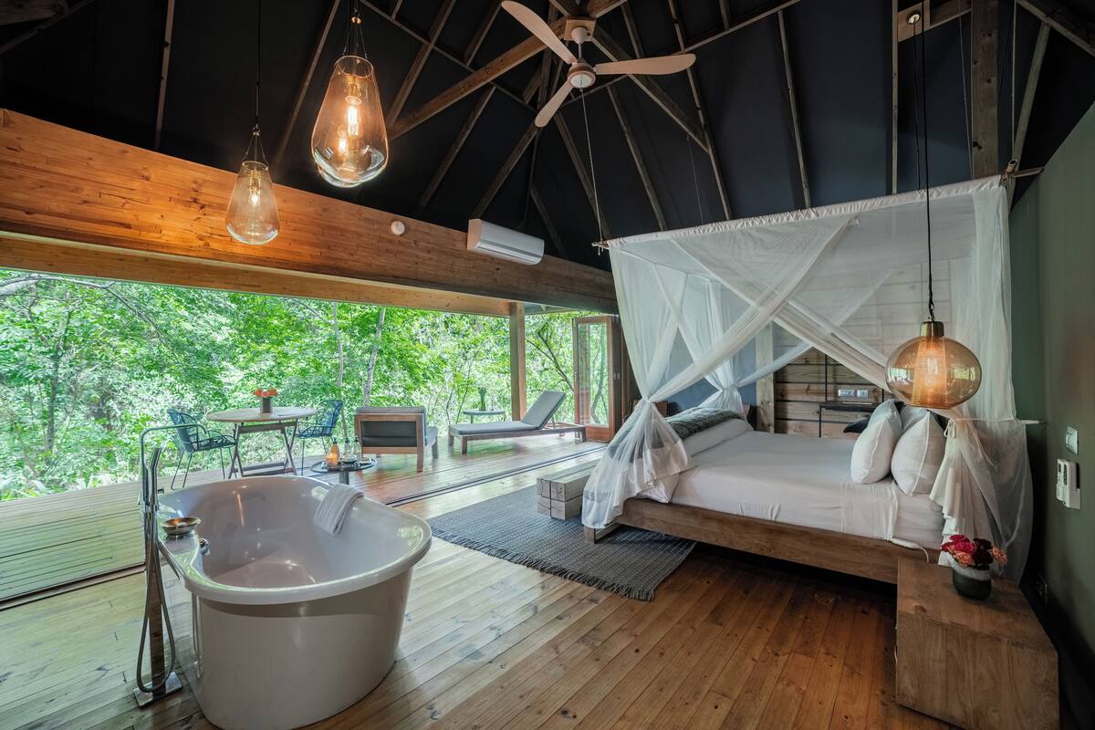 A wooden cabin with a large bathtub, a bed with a canopy, and a view of the forest.