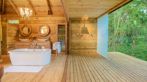 Baño de cabaña de madera con bañera independiente, pisos de madera y un ventanal con vistas a follaje.
