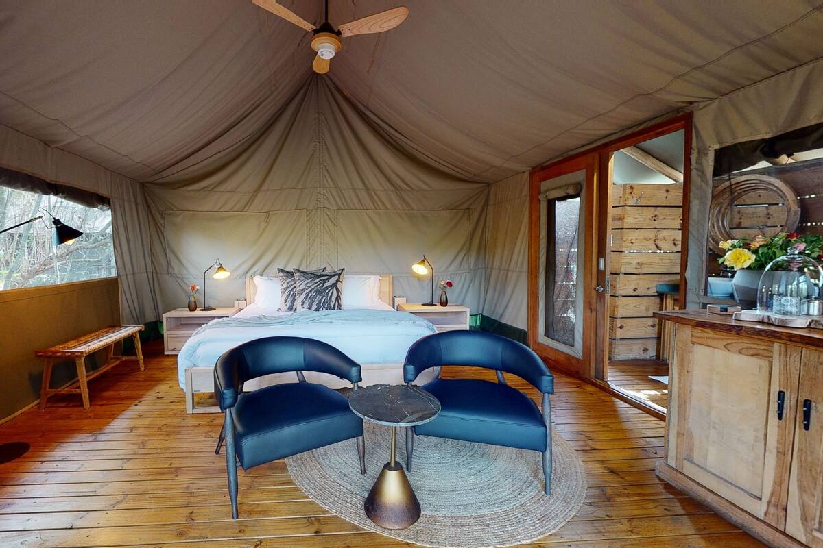 A tented room with a bed, two chairs, a small table, and a ceiling fan.