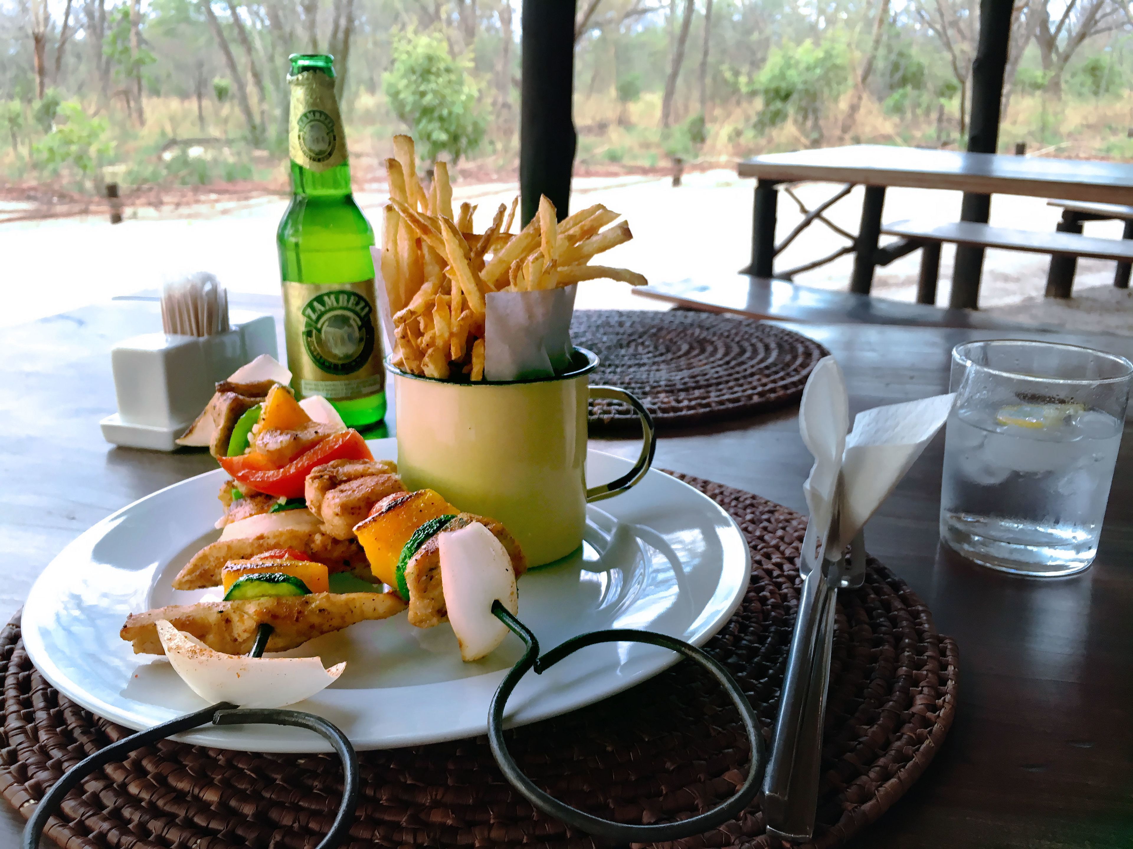 Eine Mahlzeit mit gegrilltem Gemüse, einem Sandwich, Pommes und einer Flasche Bier auf einem Tisch mit Blick auf Bäume.