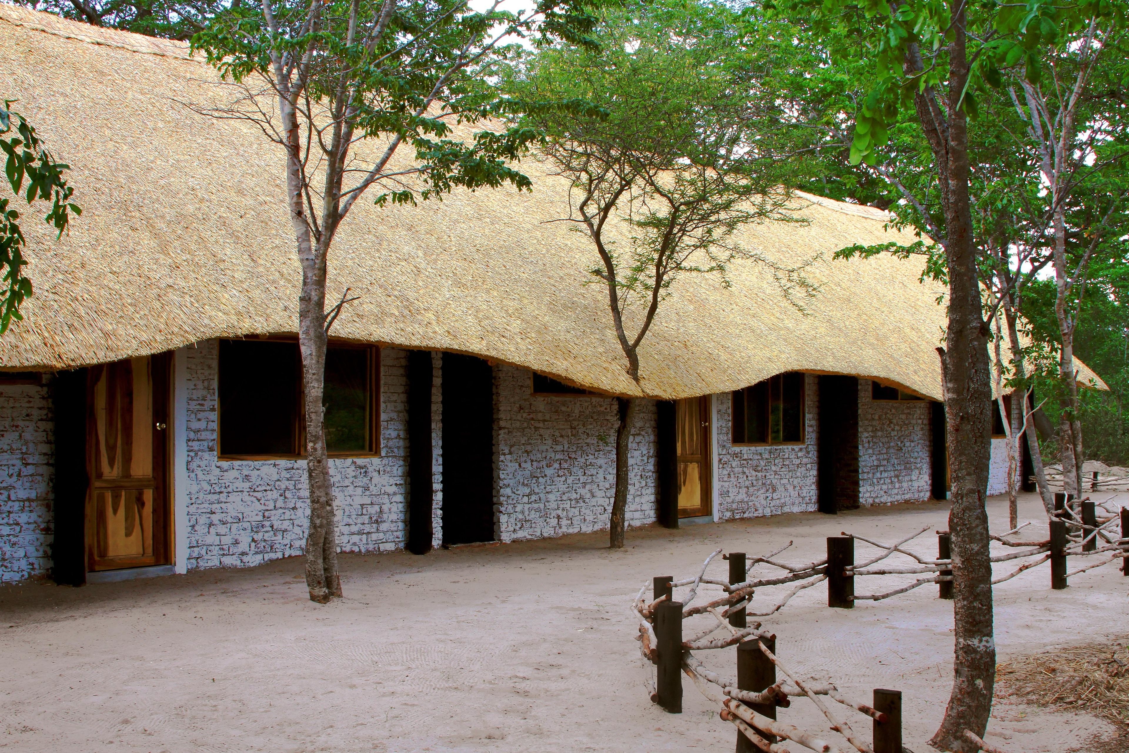 A traditional thatched-roof building with multiple rooms, surrounded by trees and a sandy ground.