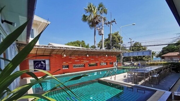 A swimming pool with a tiled deck, a red building with a sign, and palm trees in the background. - Head Rock Home (Hua Hin)