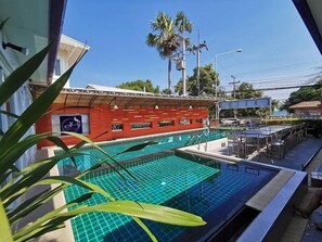 Une piscine avec une terrasse carrelée, un bâtiment rouge avec une enseigne, et des palmiers en arrière-plan.