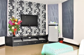 Ein modernes Wohnzimmer mit einem an der Wand mit floralem Muster befestigten Flachbildfernseher, einer schwarzen Unterhaltungsanlage mit Schubladen, einem kleinen Tisch mit grüner Tischdecke und einem Kühlschrank.
