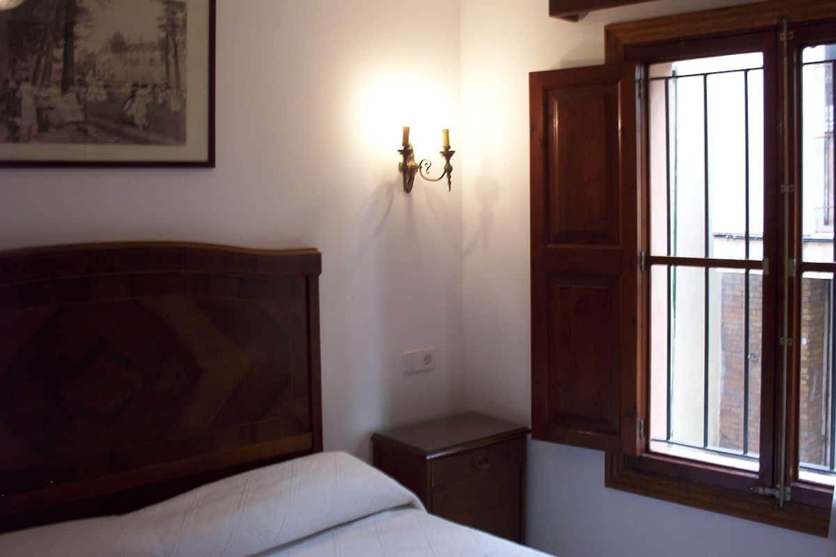 Une chambre à coucher avec une tête de lit en bois, une table de chevet, une commode, un luminaire fixé au mur et une fenêtre avec des volets en bois.