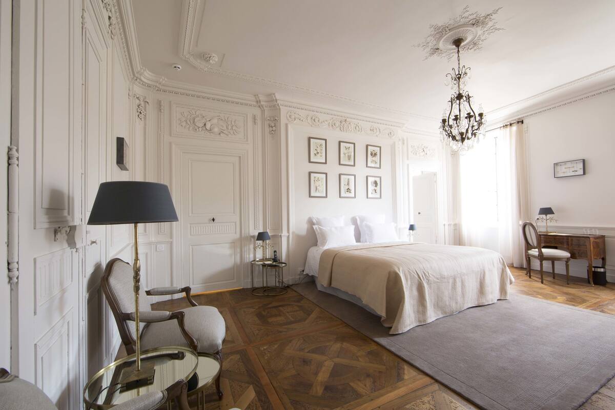 A bedroom with a large bed, a desk, a chair, a lamp, and a chandelier.