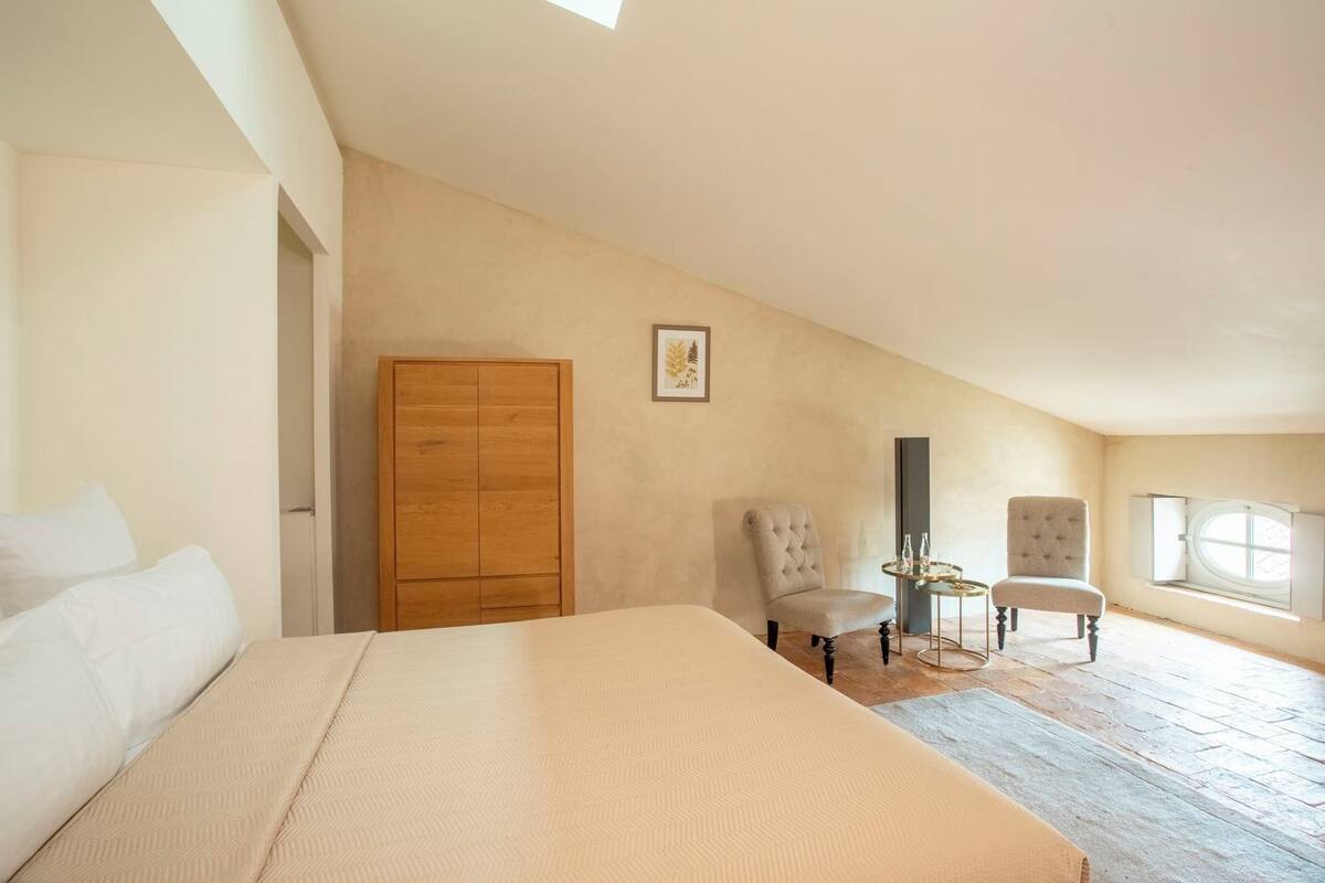 A bedroom with a bed, a wardrobe, two chairs, a small table, and a window.