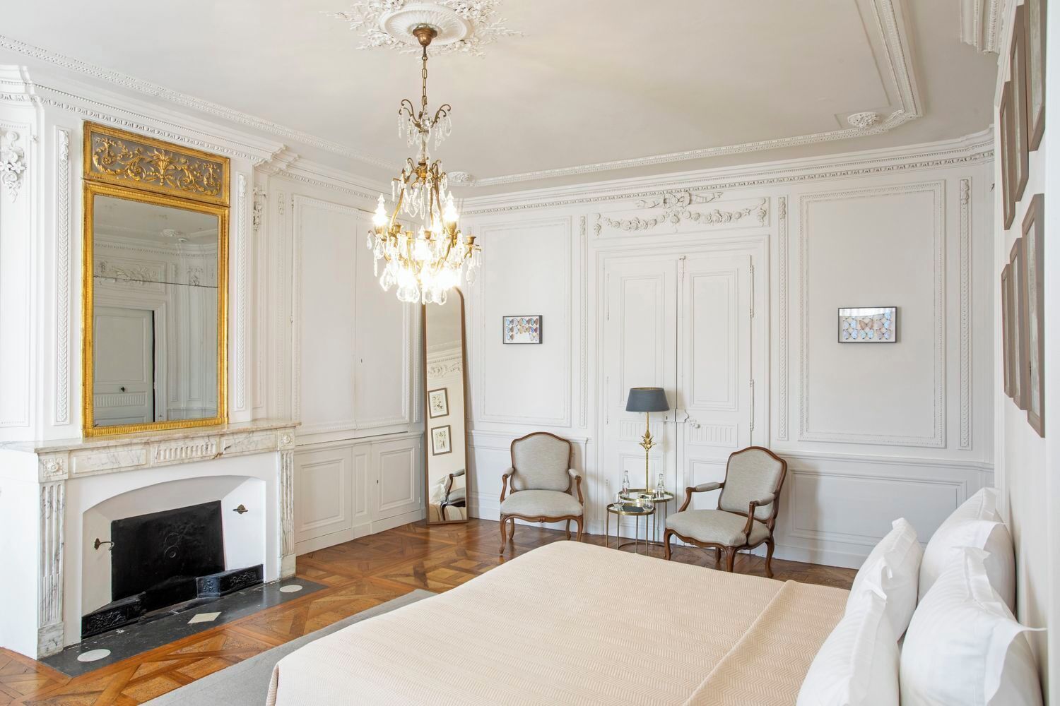 A classic, elegantly furnished bedroom with a large bed, two chairs, a fireplace, and a chandelier.