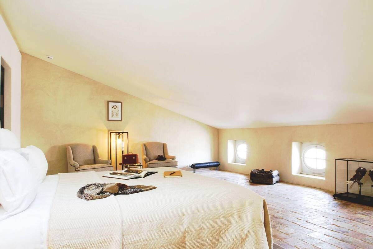 A spacious attic bedroom with a large bed, two armchairs, a desk with a lamp, and a window.