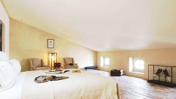 A spacious attic bedroom with a large bed, two armchairs, a desk with a lamp, and a window.