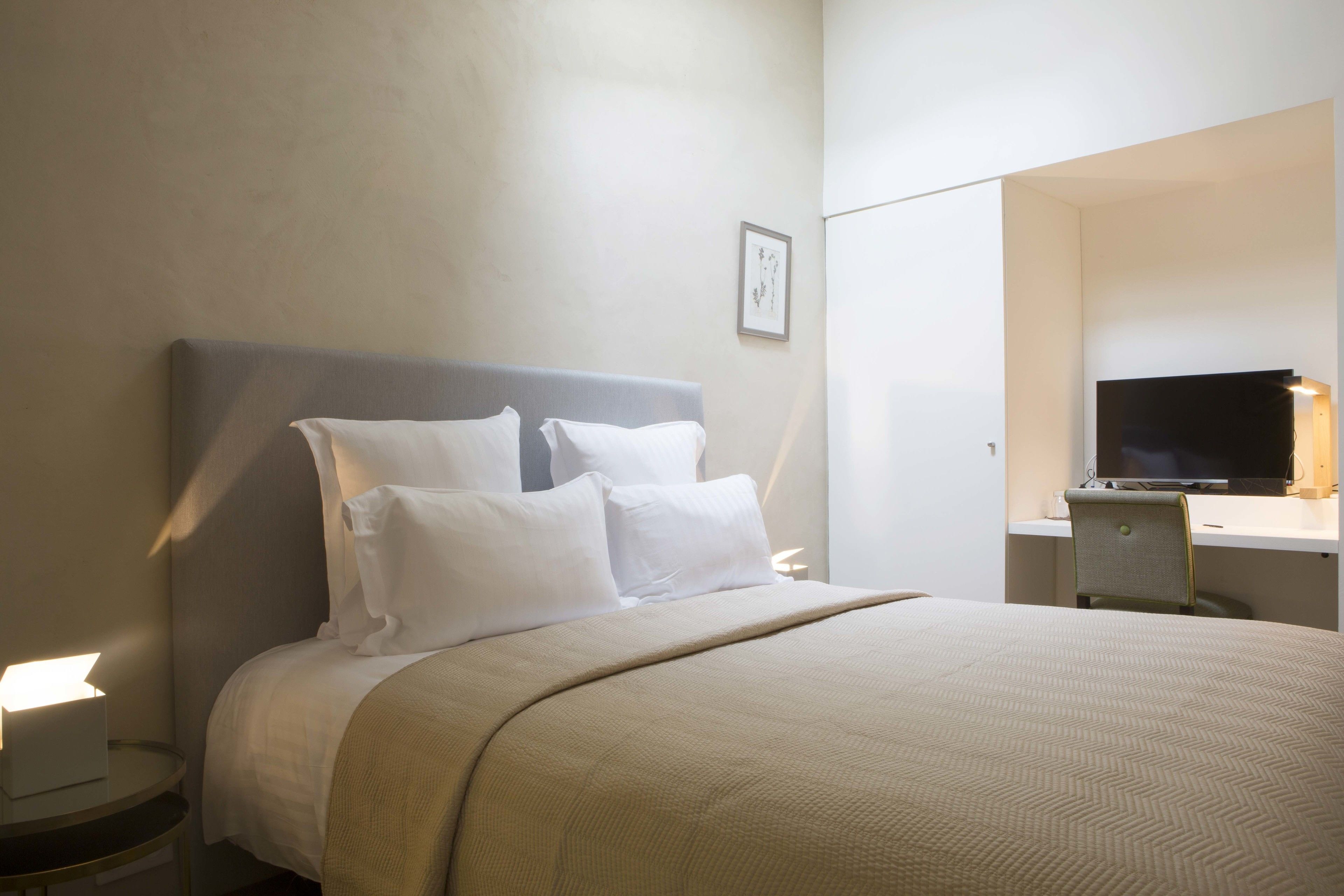 A neatly made bed with white linens, a bedside table with a lamp, and a desk with a computer and a chair.